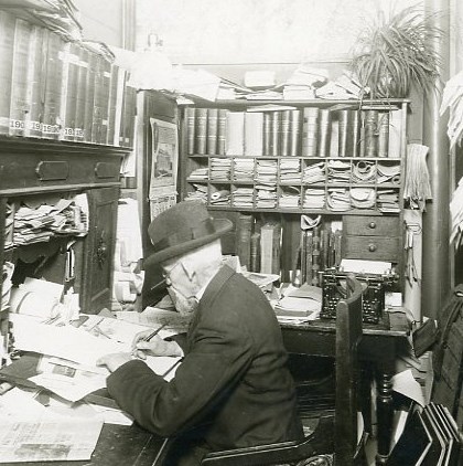 A black and white image of a man working at a desk filled with papers and books, c. 1910. 