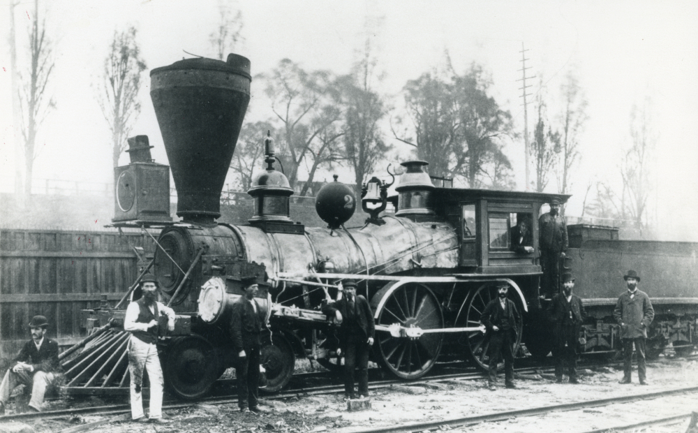 A black and white photo of an old steam-engine with 9 men standing in and around it. 