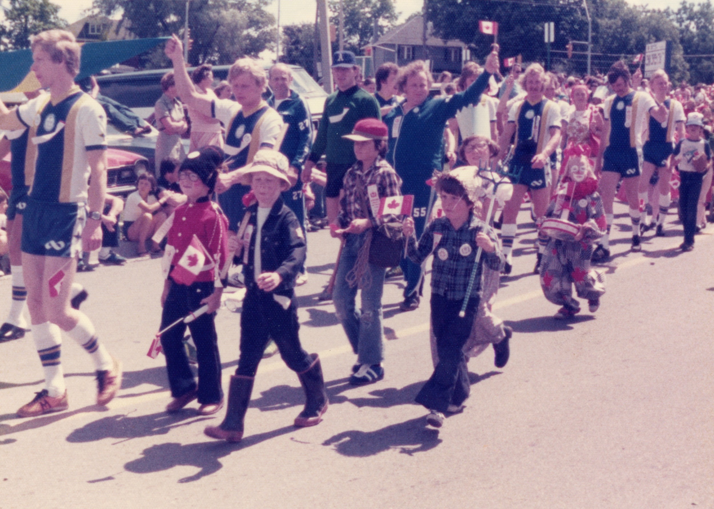 A colour photo of kids of various ages walking down the road participating in a Canada Day parade, 1977