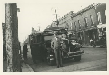 Black and white photograph a 1920s street scene showing a school bus parked on the site of the road with a man standing in front of it. 