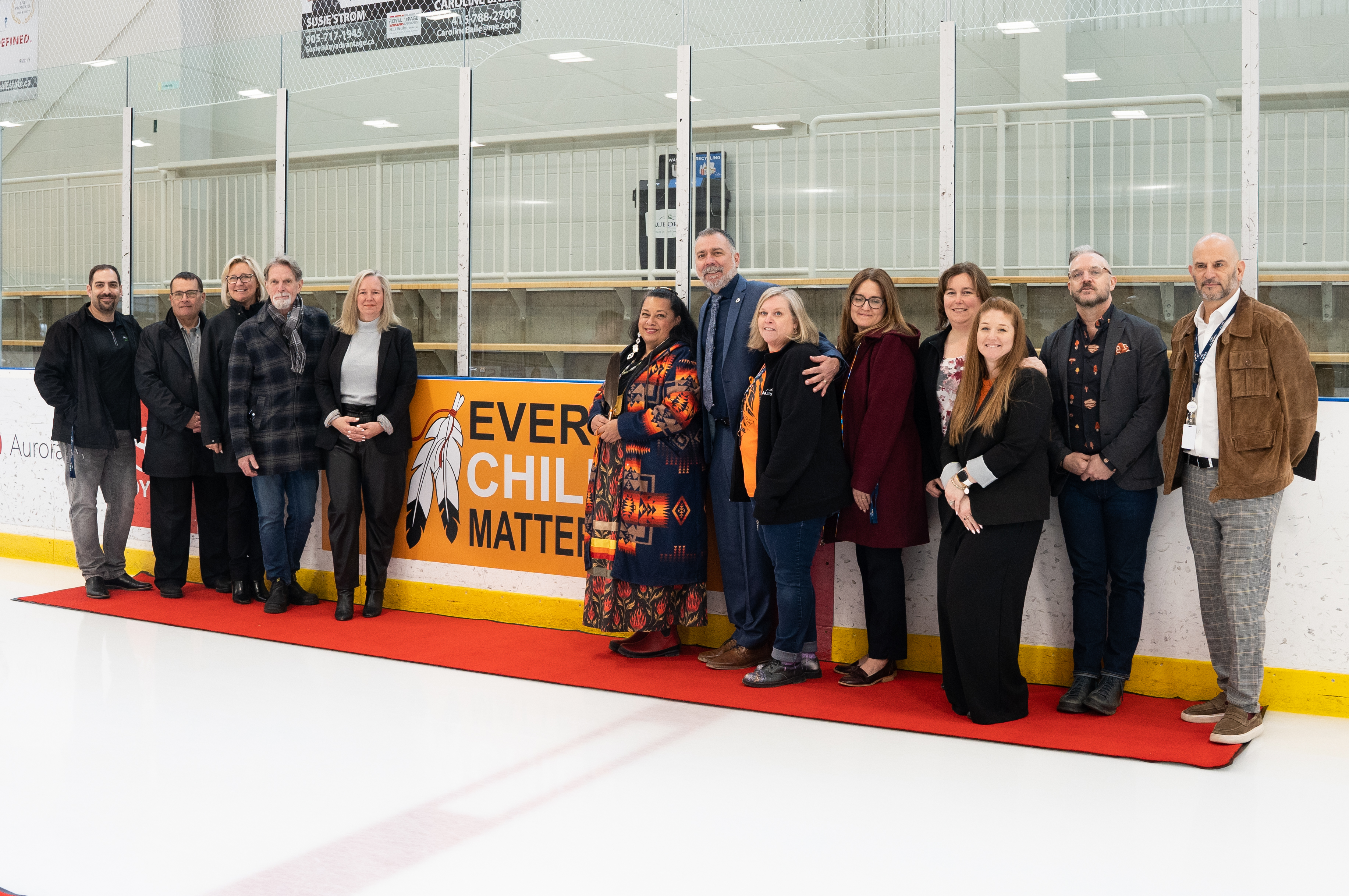 Mayor, council, staff and Kim Wheatley, Anishinaabe Ojibway Grandmother in front of Every Child Matters Rink Board