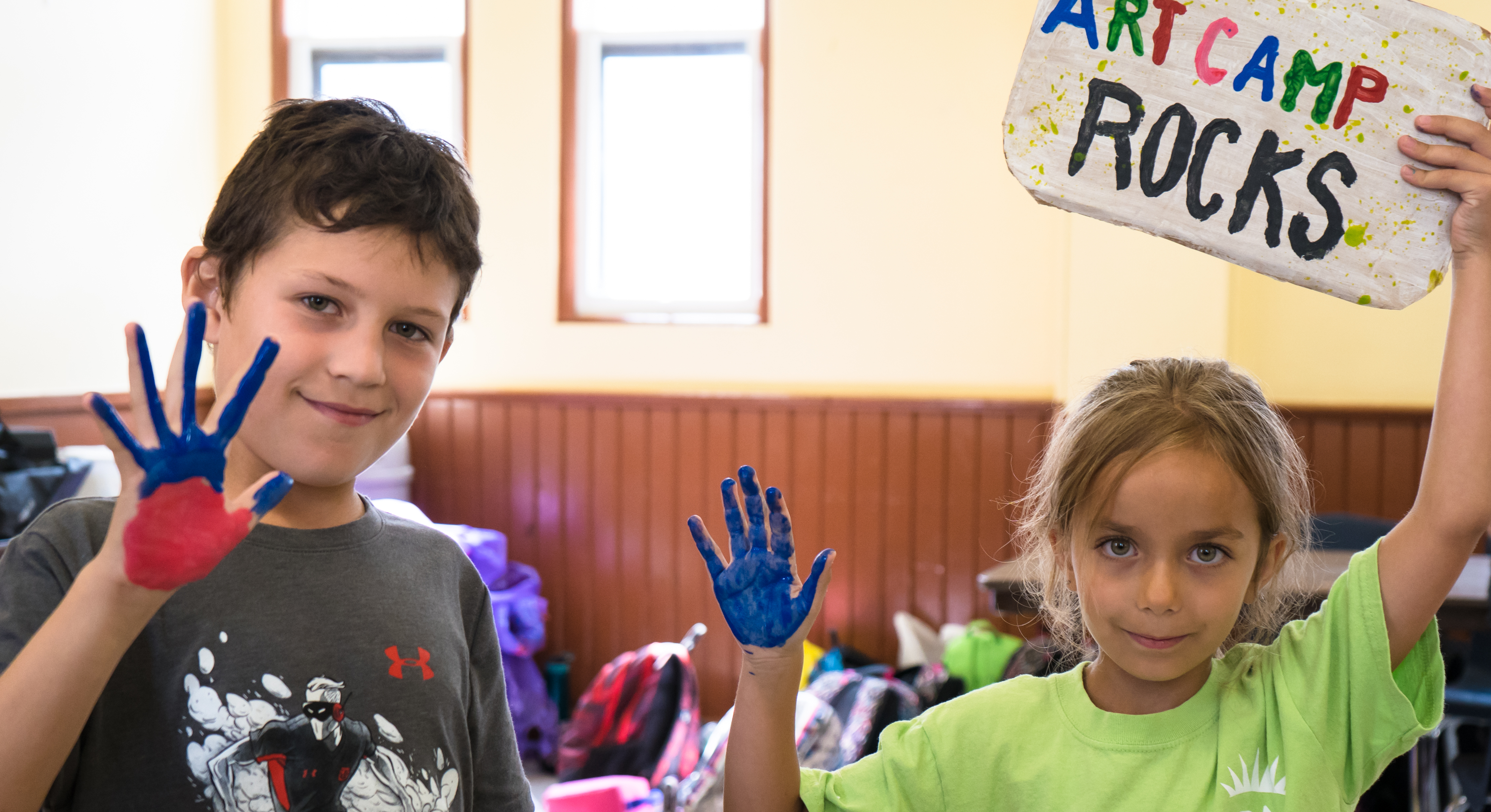 Boy and girl with hands covered in blue paint holding a sign that says "Art Camp Rocks".