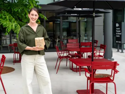 Meagan in front of the cafe, with red chairs and a tree, holding a bag of coffee and smiling