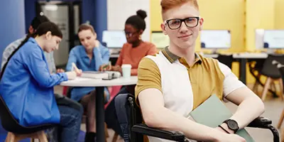 Young person in a wheelchair, smiling, with more people seated behind him in the distance