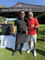 Two men on a lawn, in front of a large tent, one holding a skewer
