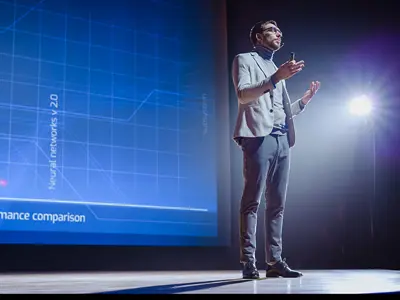 Man on stage presenting, with a big blue screen behind him