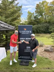 Two men on green grass in front of a sign and holding a drink each