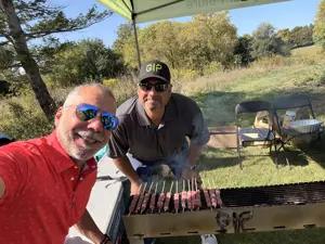 Two men posing for a selfie, one tending to a grill