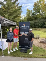 Two men and a woman, beside a popup promo sign, two holding up drinks