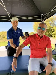 The mayor sitting on a bench and another one holding his arm, both under a tent