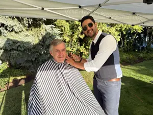 Two men under a tent, one putting a barber's apron on the other