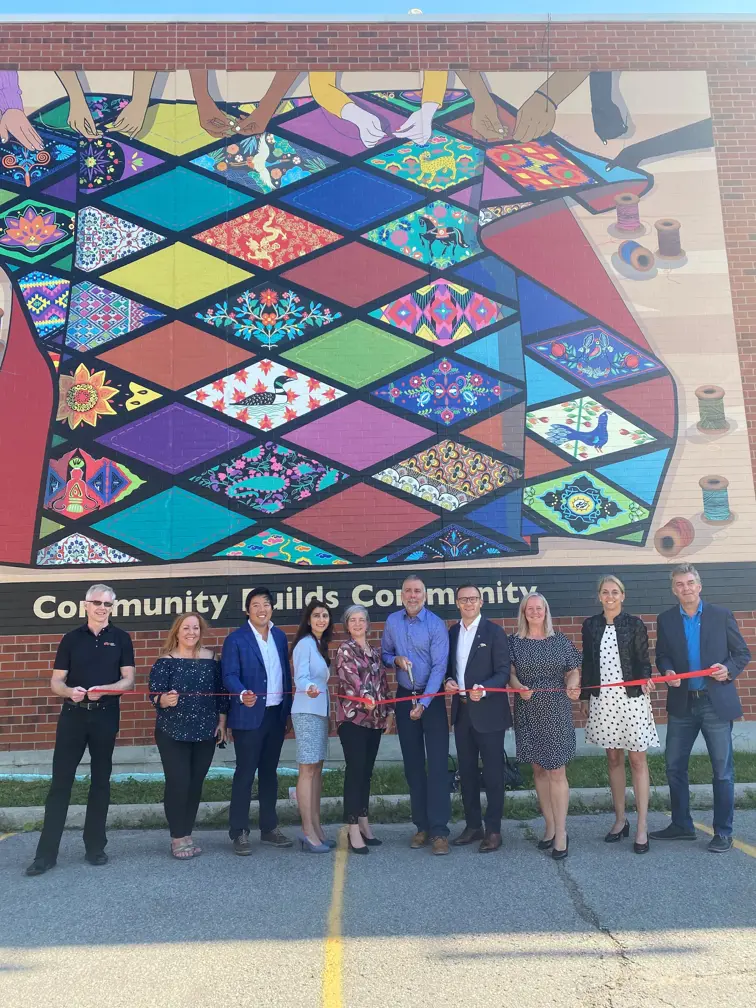 Group of people in front of a building and mural, all holding a ribbon and one person holding large scissors
